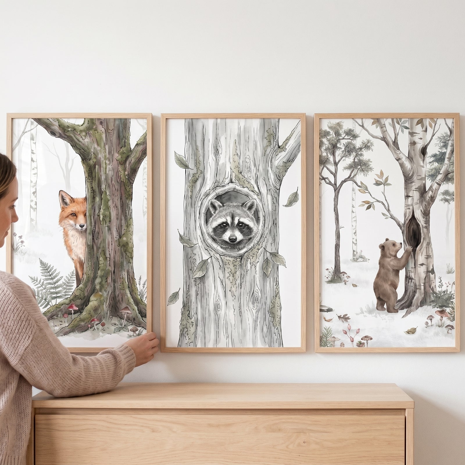 Woman adjusts a neutral woodland animals wall art set of 3 with a fox, raccoon, and bear over a wooden dresser.