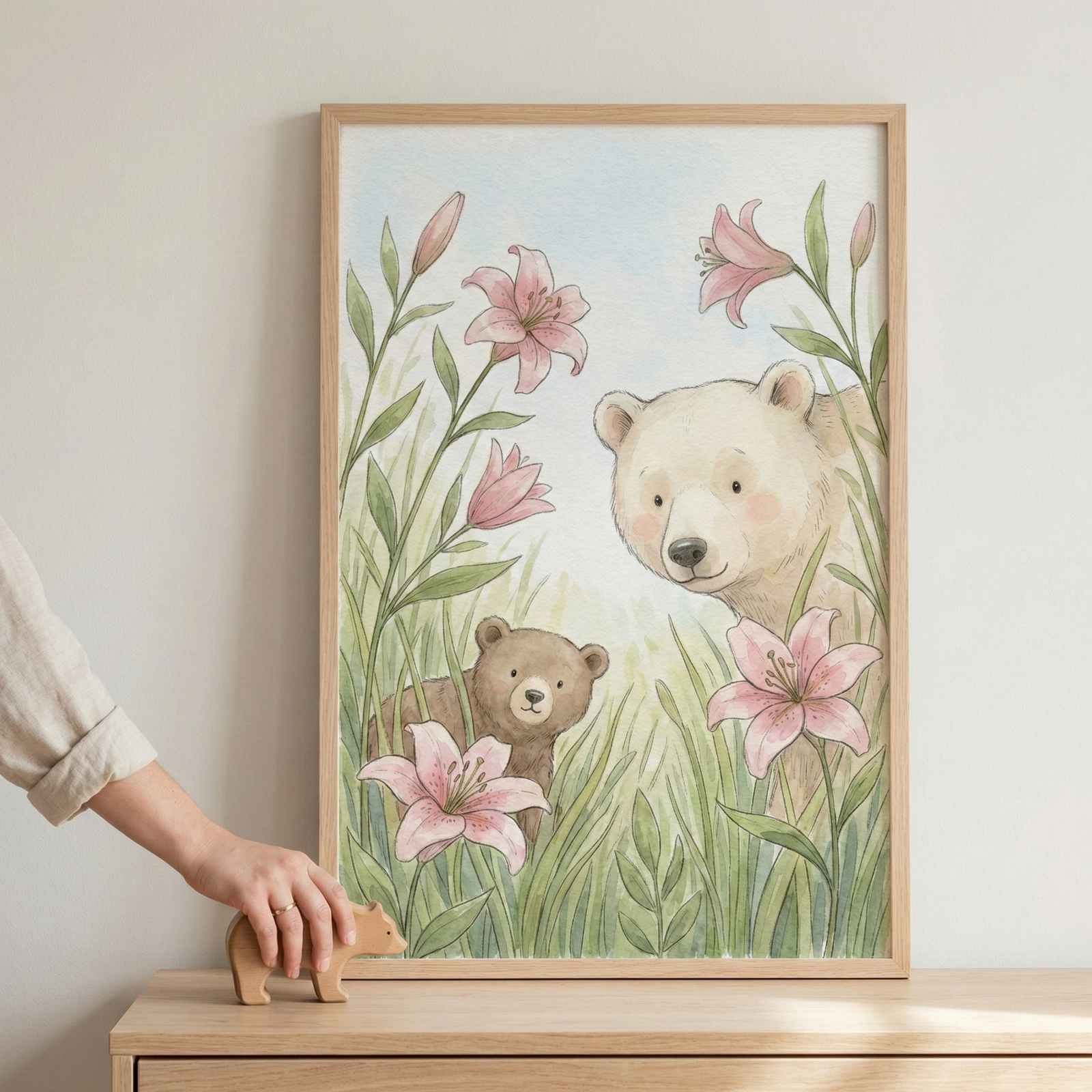 A framed illustration depicts a charming bear and cub peeking through tall grass and pink lilies, set against a soft blue sky. A hand is placing a wooden bear figurine on the wooden surface below the artwork.