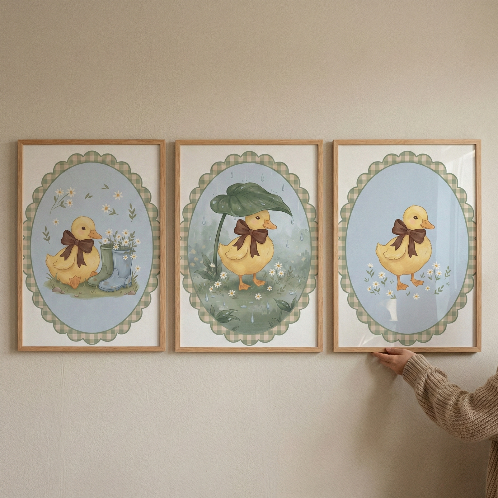 Three wooden frames hang on a neutral wall, each showcasing a charming illustration of a yellow duckling adorned with a brown bow. Each scene is encased in a scalloped oval border with a green and white checkered pattern. The left frame shows a duckling sitting beside blue boots filled with flowers. The middle frame features a duckling using a large leaf as an umbrella. The right frame depicts a duckling standing in a field of small white blossoms. A hand in a beige sweater touches the right frame.