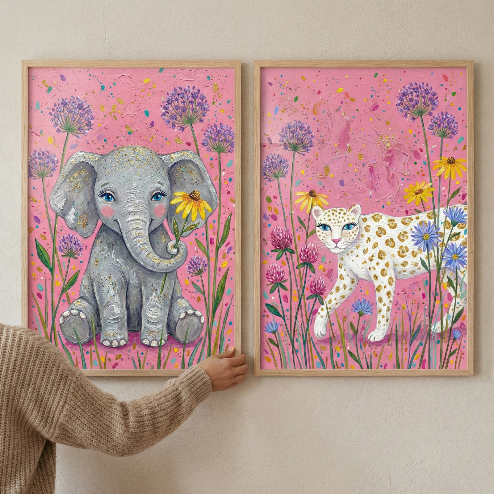 Two rectangular, light wood-framed art pieces hang side-by-side on a neutral wall. A person's arm, clad in a beige cable-knit sweater, is visible as their hand adjusts the left frame. Both illustrations share a whimsical style with textured, pink backgrounds speckled with colorful dots. The left piece features a charming gray elephant with blue eyes holding a yellow blossom. 