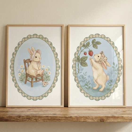Two framed illustrations of a small beige rabbit rest on a wooden shelf against a neutral wall. Each artwork features a rabbit in a floral bonnet within a scalloped, checkered oval border. On the left, the rabbit sits on a tiny wooden chair with a teacup and daisies nearby. On the right, the rabbit stands on its hind legs to reach a red strawberry on a vine. The soft, gentle style uses a palette of light blue, green, and cream.