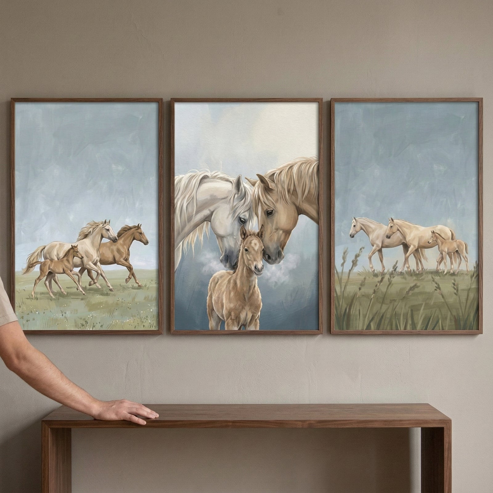 A triptych of three vertical framed illustrations depicting horses hangs on a neutral beige wall. Each piece is housed in a simple dark wood frame and features a soft, ethereal style with a palette of blues, greens, and warm browns.

The leftmost illustration shows three horses running across a green meadow. A white horse and a brown horse are in full stride, accompanied by a smaller brown foal running beside them against a pale, cloudy sky.
