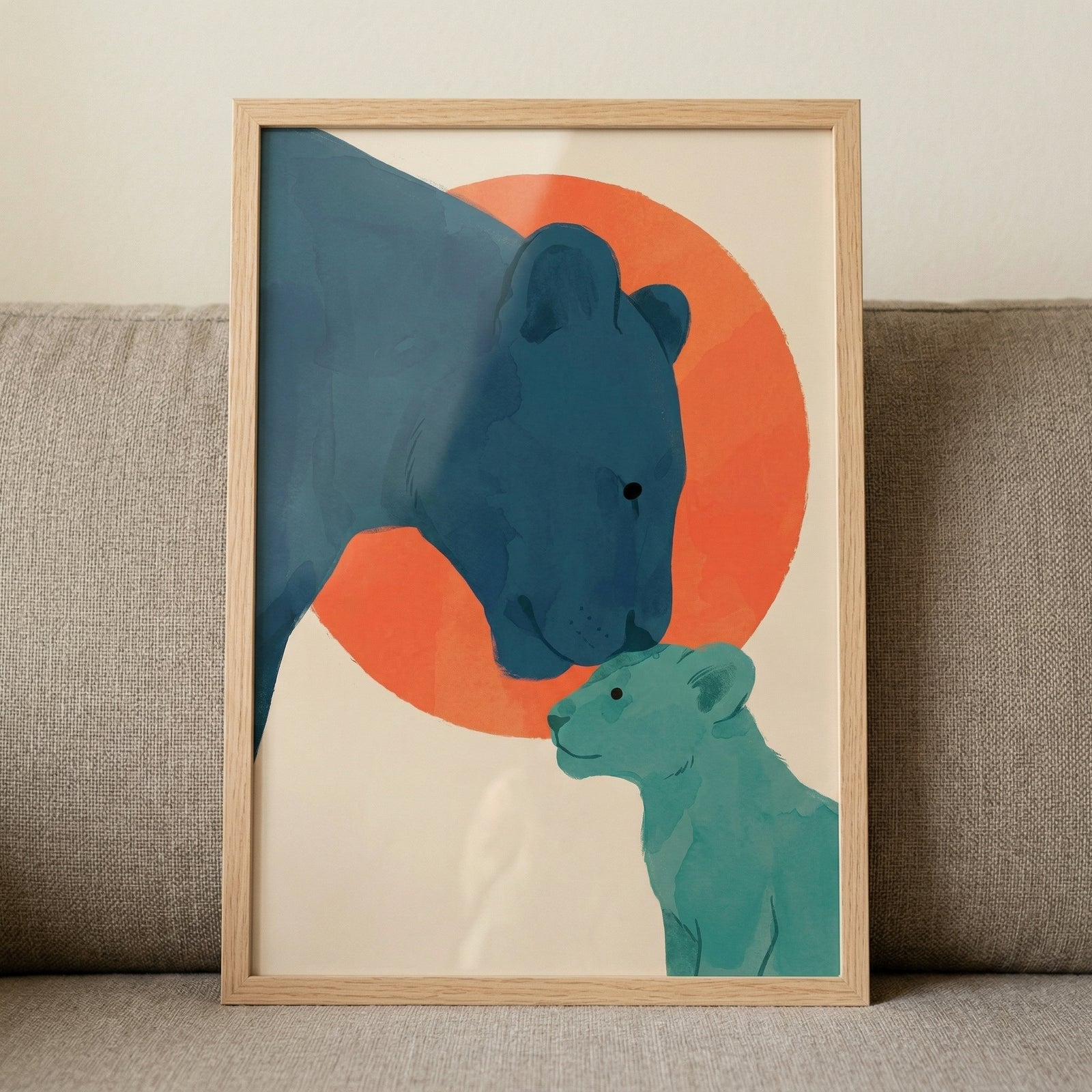 A framed modern graphic illustration depicting a mother big cat and her cub in a tender moment. The larger feline is rendered in deep blue shades, while the smaller cub is a soft mint green. Both figures are shown in profile against a cream-colored background featuring a large, vibrant orange circular sun. The artwork uses a minimalist, flat color style and is housed in a light wood frame, propped against a textured grey couch.