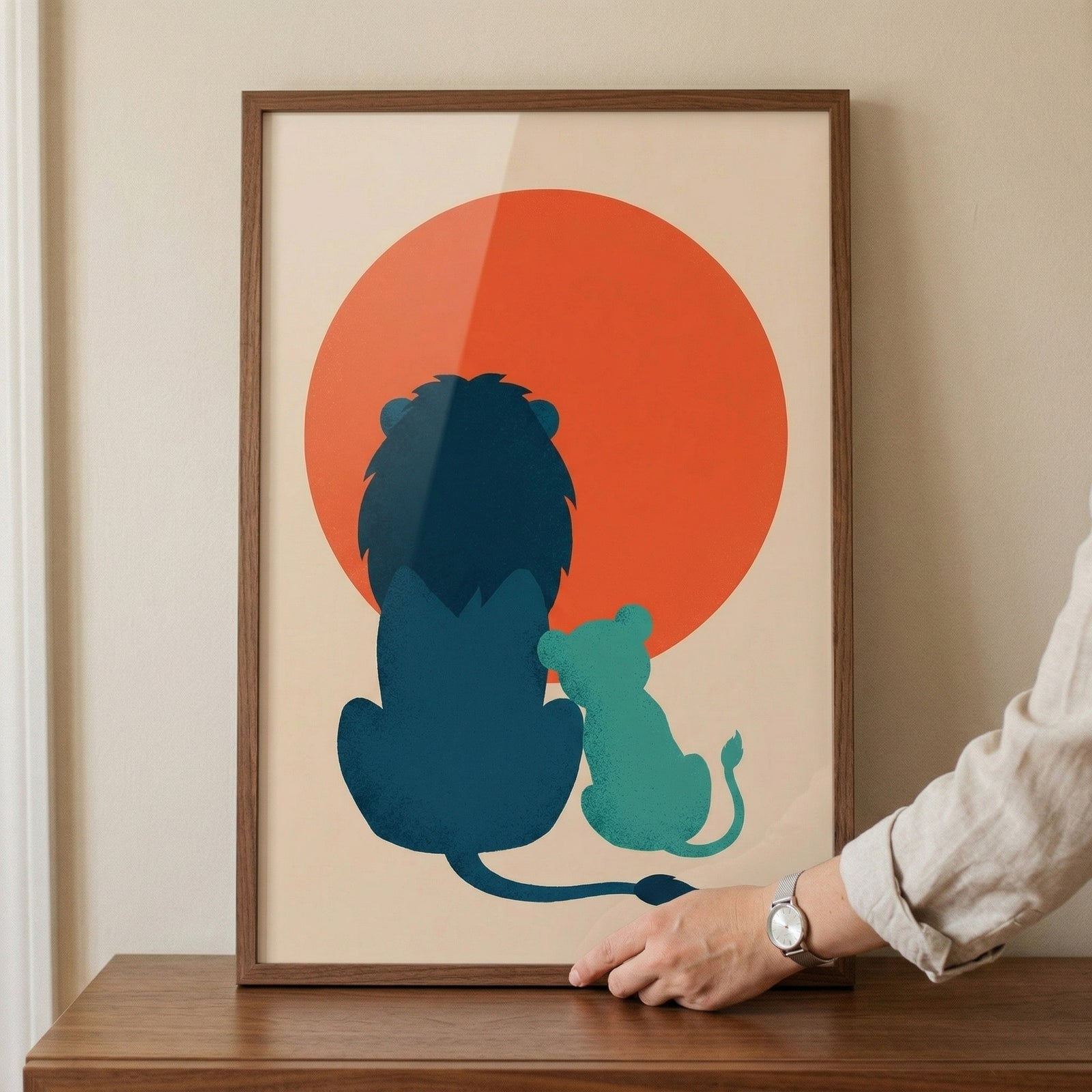 Minimalist framed artwork of a dark blue lion and smaller cub sitting side by side in front of a large warm red sun. A hand adjusts the frame on a wooden surface highlighting the print as a calming decorative piece for a nursery or child’s room.