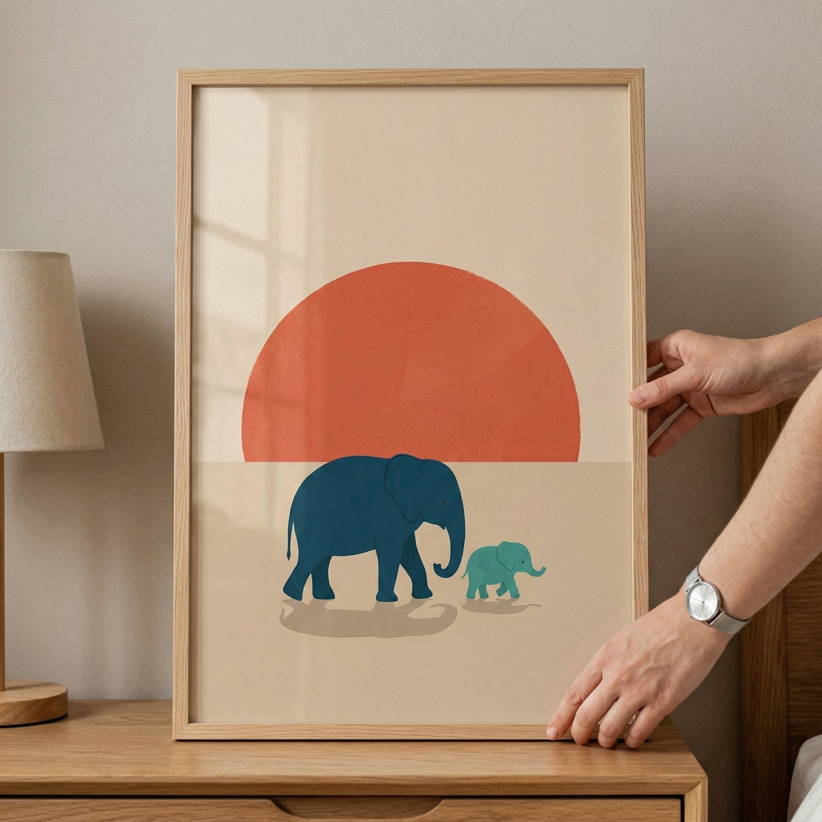 Framed illustration of a large blue elephant walking beside a smaller green baby elephant in front of a bold red sun, placed on a wooden dresser. The simple shapes and warm tones create a calm and playful focal point for a child’s room.