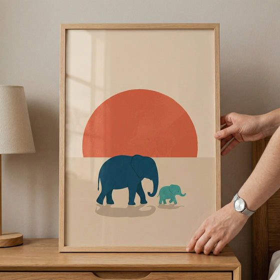 Framed illustration of a large blue elephant walking beside a smaller green baby elephant in front of a bold red sun, placed on a wooden dresser. The simple shapes and warm tones create a calm and playful focal point for a child’s room.
