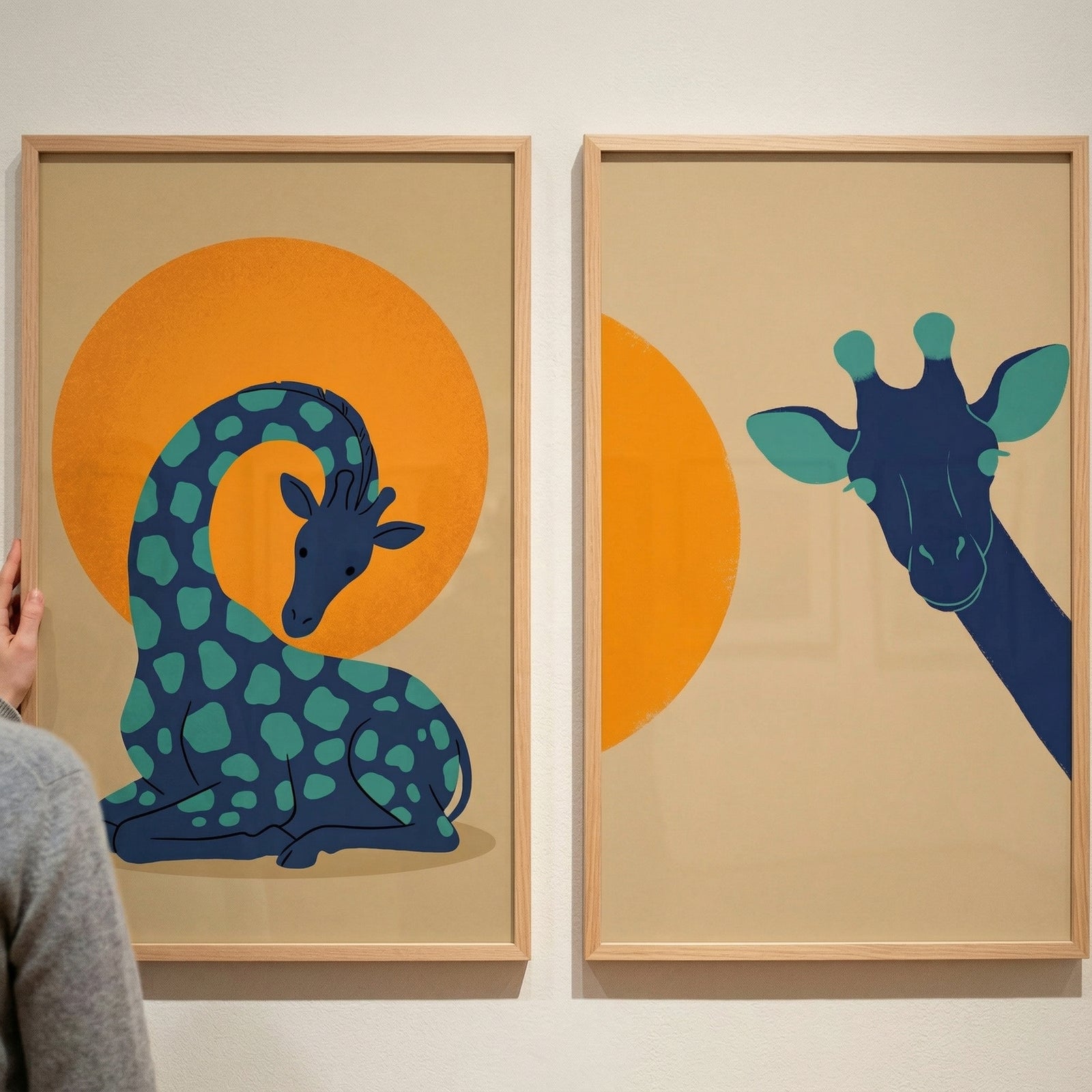Two light wood frames containing graphic illustrations are displayed side-by-side on a neutral-colored wall. The illustration on the left features a stylized dark blue long-necked animal with teal spots, resting in a curled position in front of a large, solid orange circle. 