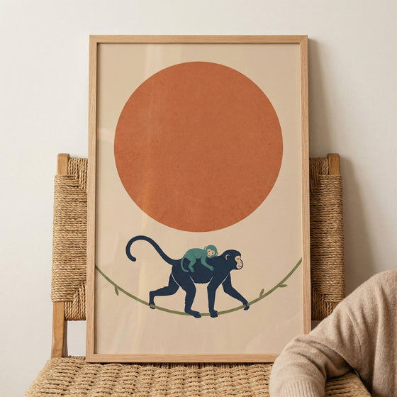 Framed illustration of a monkey carrying a baby on its back while walking along a branch beneath a large orange sun. The artwork rests on a woven chair, adding a warm and playful jungle inspired decor touch.
