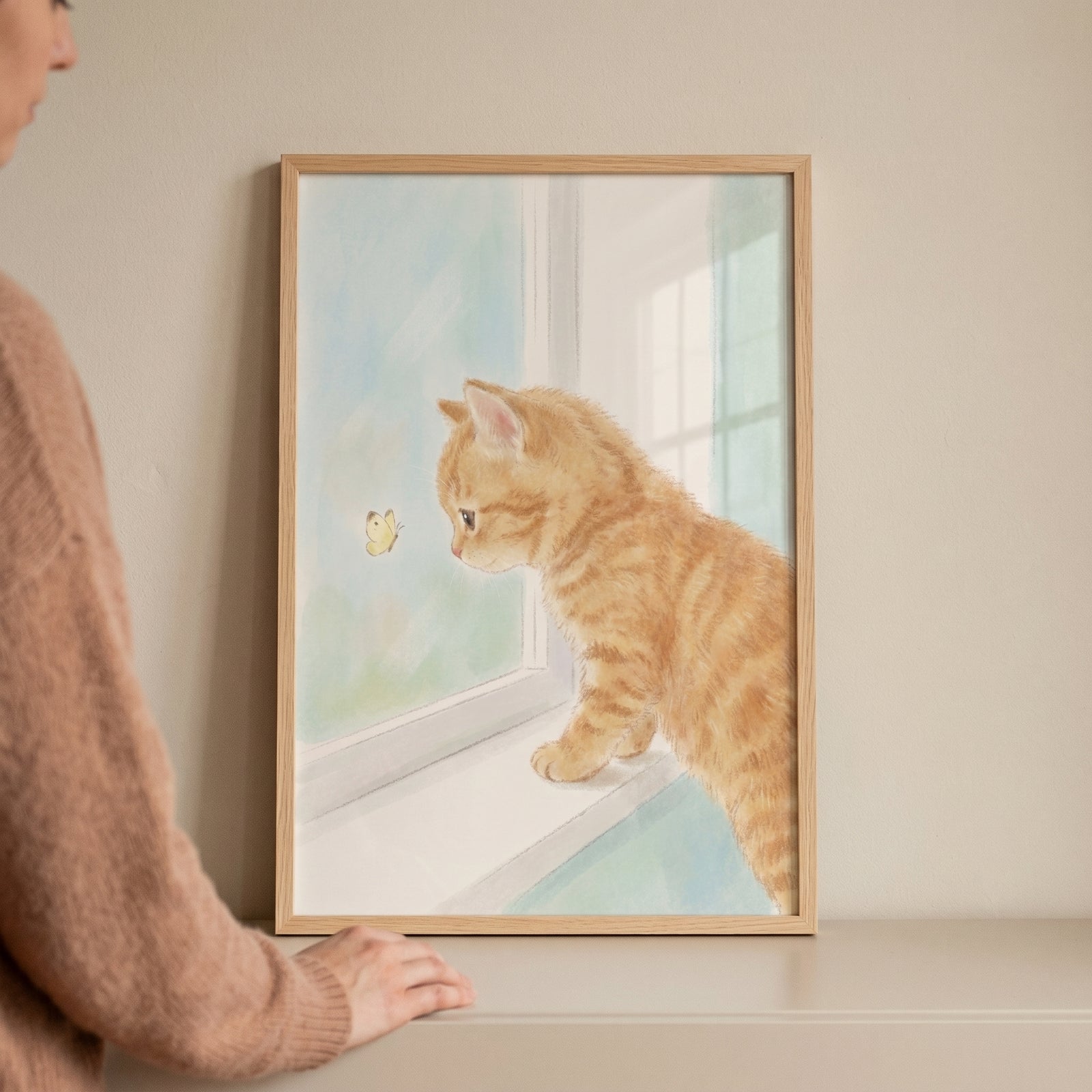 A person touches framed orange tabby kitten wall art of a kitten watching a butterfly from a window ledge.