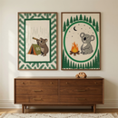 Two vertical framed illustrations hang side-by-side on a light wall. The left print shows a brown koala character using a telescope next to a small tent marked "01" against a rainy backdrop, bordered by a green evergreen needle pattern. The right print features a grey koala character sitting by a campfire under a moonlit sky, enclosed in a circle and surrounded by forest silhouettes. Below the art sits a mid-century modern wooden dresser with six drawers and brass hardware.