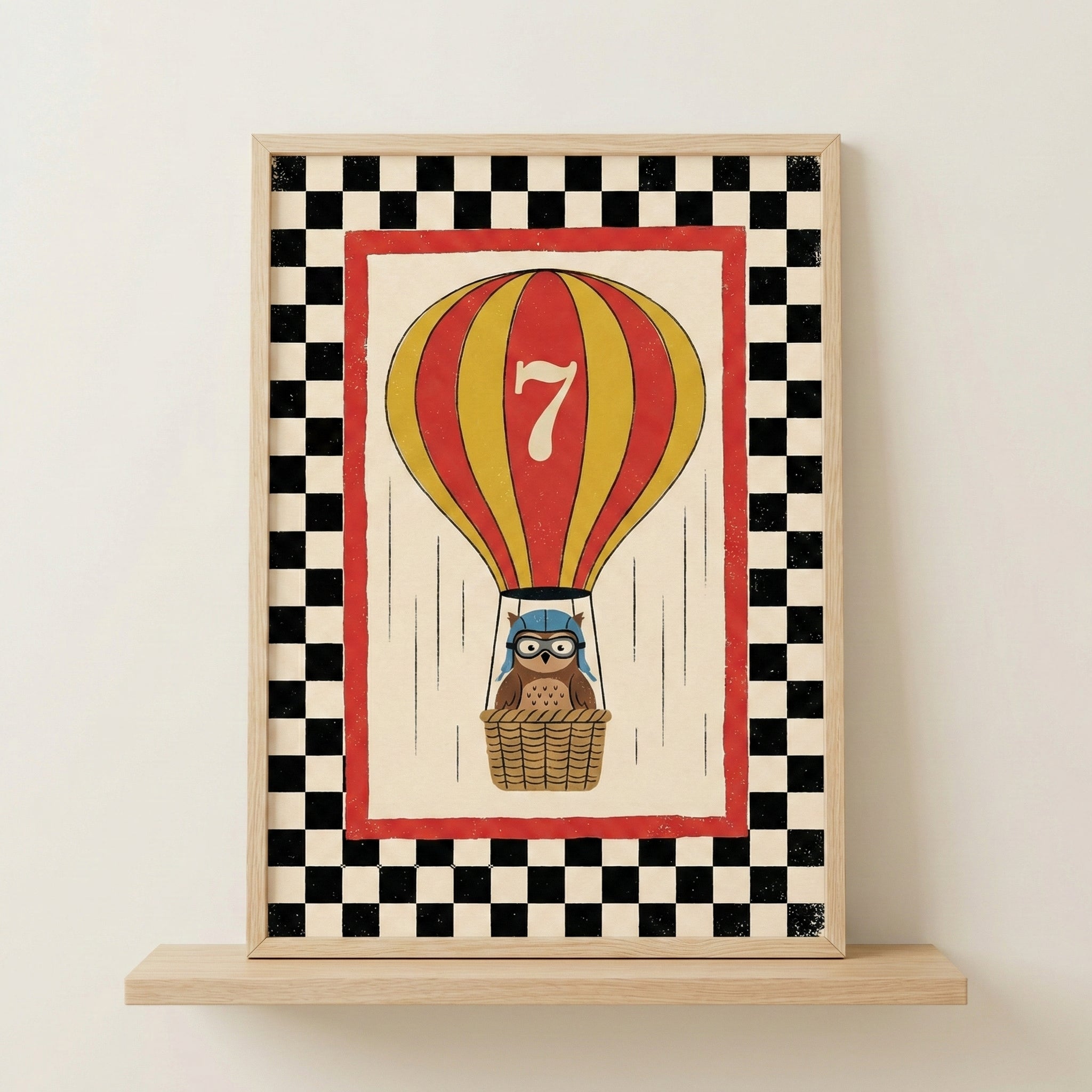 A framed wall art print features a whimsical illustration of a red and yellow striped hot air balloon with a large white number seven on its side. In the woven basket below, a small brown owl wears a blue aviator cap and goggles. The central image is surrounded by a solid red border, which is enclosed by a bold black and white checkerboard pattern. The artwork is presented in a light-colored wood frame and sits on a matching wooden shelf against a neutral wall.