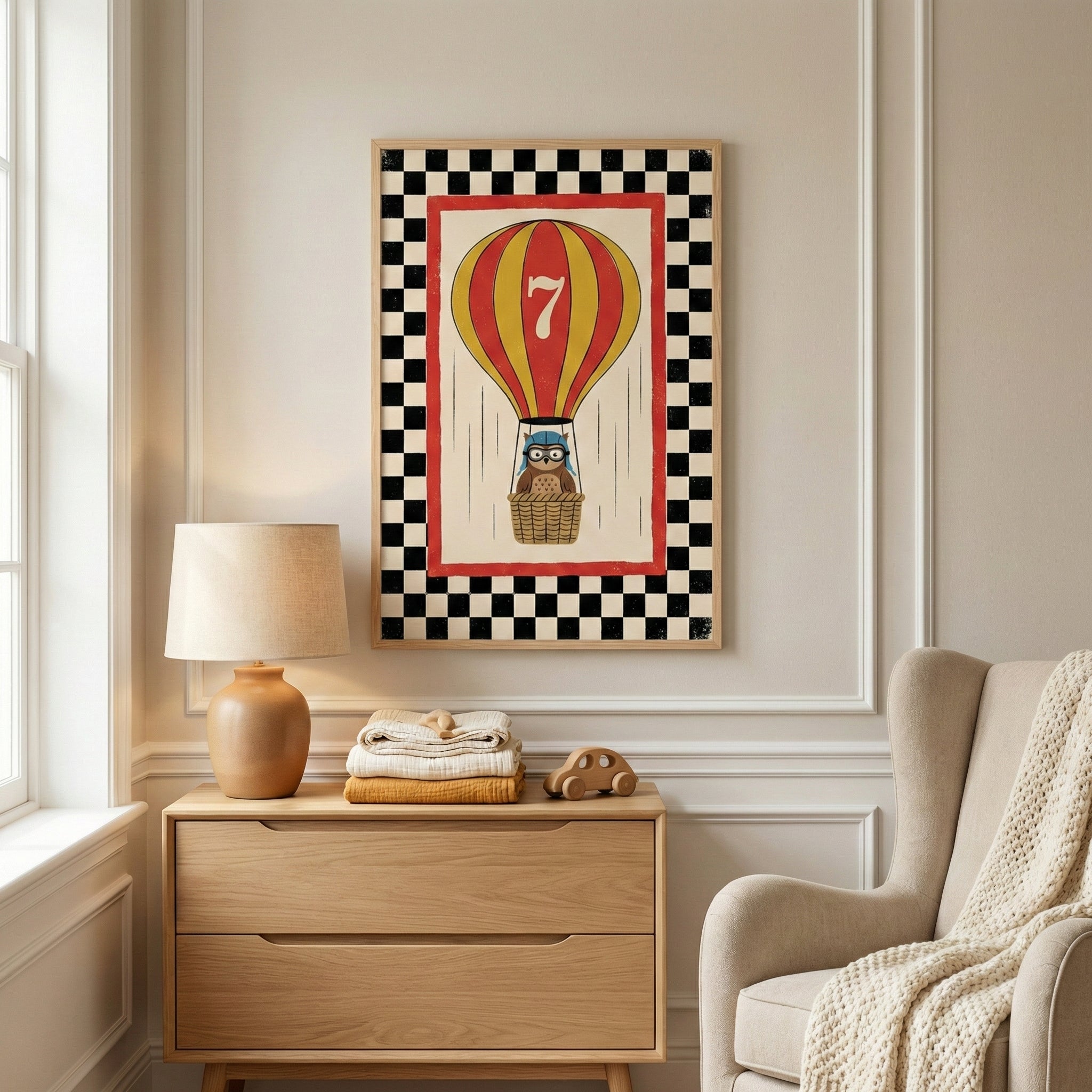 A vertical framed artwork hangs on a light-colored wall with classic molding in a cozy room. The art shows a red and yellow striped hot air balloon with a large number 7 on it, carrying a small owl wearing aviator goggles in its basket. The piece is bordered by a black and white checkered pattern and a red rectangular frame. Below the art sits a light wood dresser holding a tan lamp, a stack of folded blankets, and a small wooden toy car. 