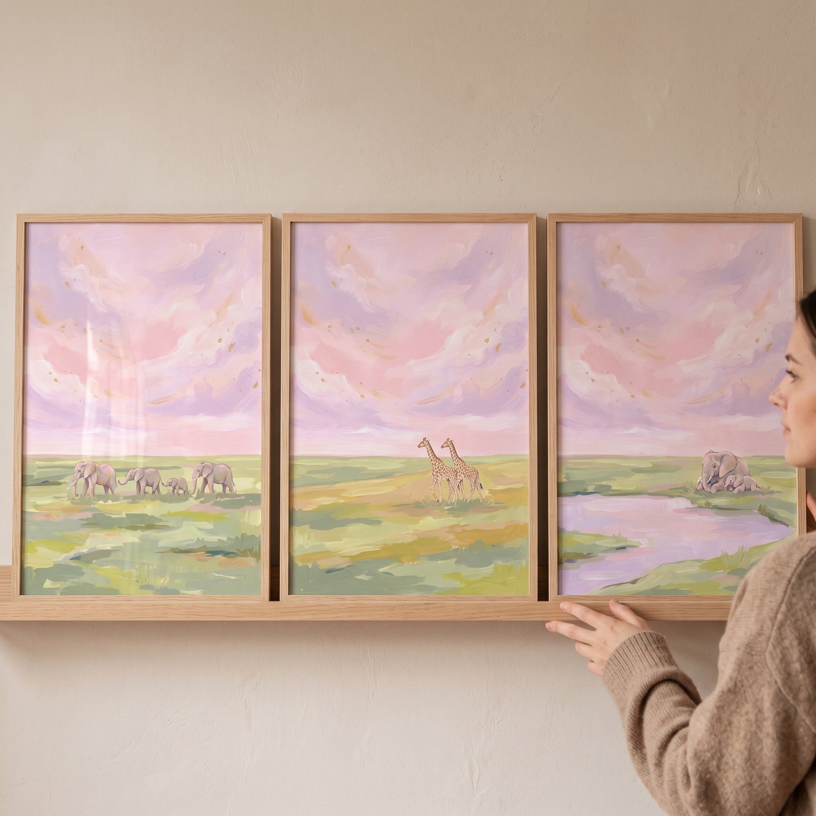 Woman places a painterly blush safari animals wall art set of 3 with giraffes and elephants on a wooden shelf.