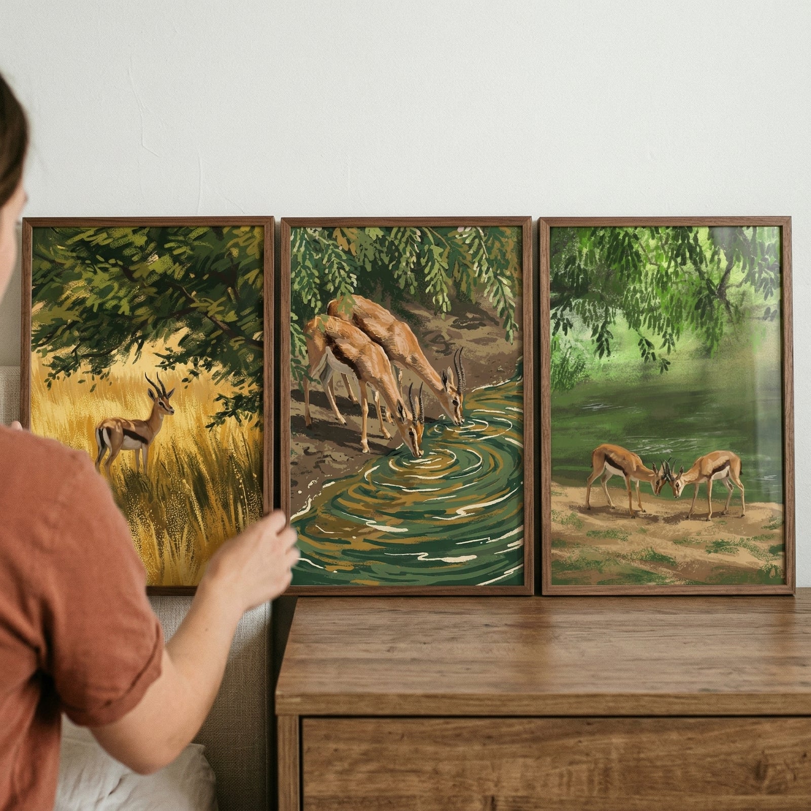 A hand touches a painterly green savanna gazelle wall art set of 3 featuring wild animals by trees and a water source.
