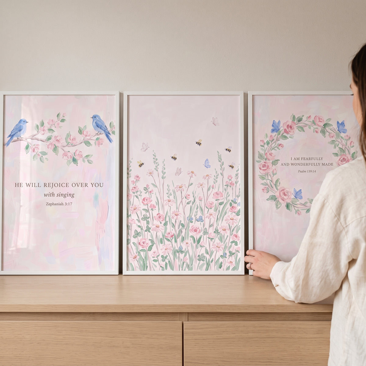 A woman arranges a painterly pink rose botanical artwork set of 3 featuring birds and Bible verses on a wood dresser.