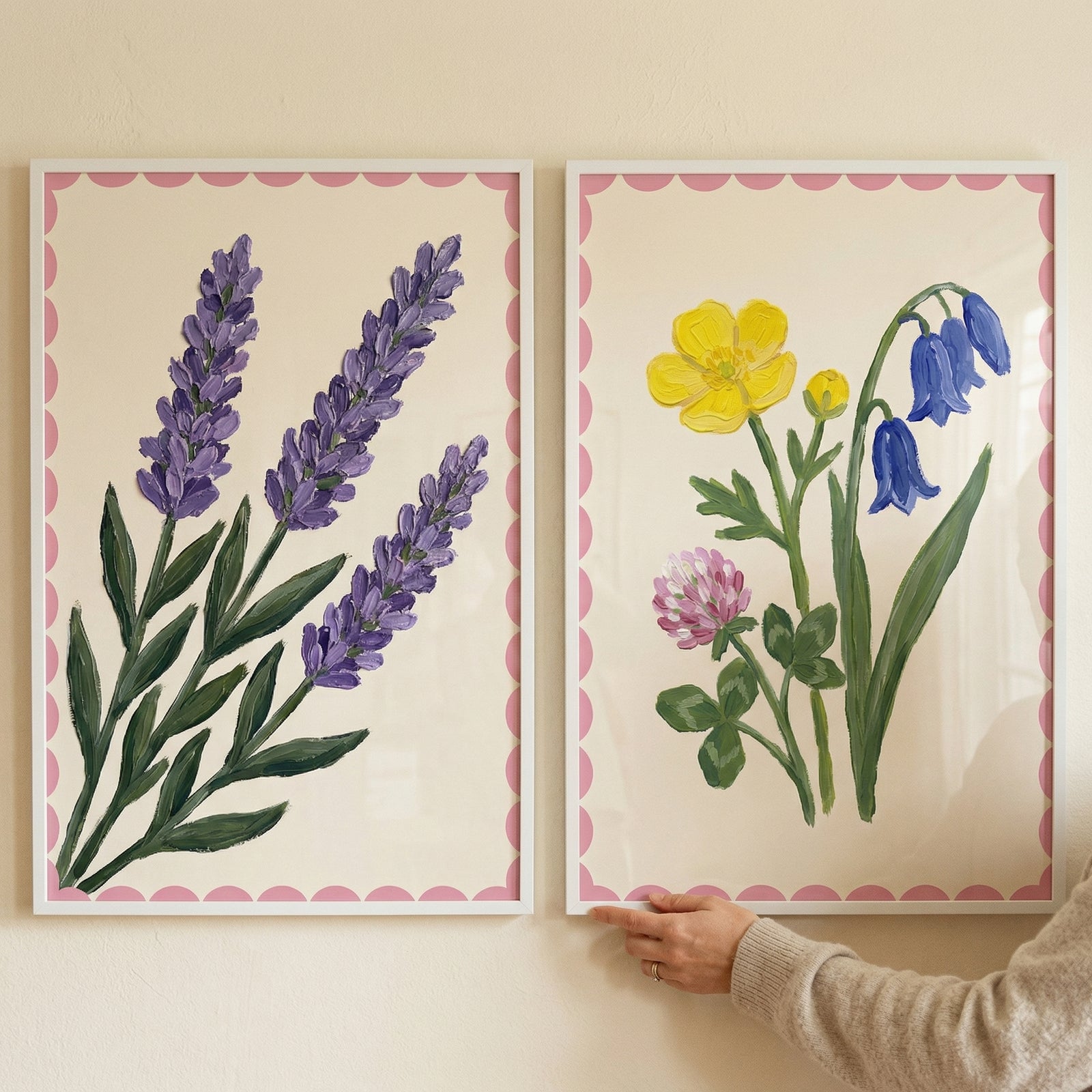 Two framed botanical prints with pink scalloped borders hang on a neutral wall, one showing purple lavender stems and the other featuring a mix of yellow, blue, and pink flowers. A hand gently adjusts the right frame, suggesting the artwork being styled or arranged on the wall.