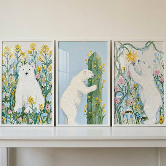 A triptych of white-framed artworks displayed on a white shelf. Each illustration features a white polar bear in a whimsical, colorful setting. On the left, a bear sits among tall yellow and blue flowers. In the center, a bear stands and leans against a tall green pillar covered in small yellow blossoms. On the right, a bear reaches upward towards a golden star among flowering vines. The illustrations have a soft, textured appearance with visible artistic strokes.
