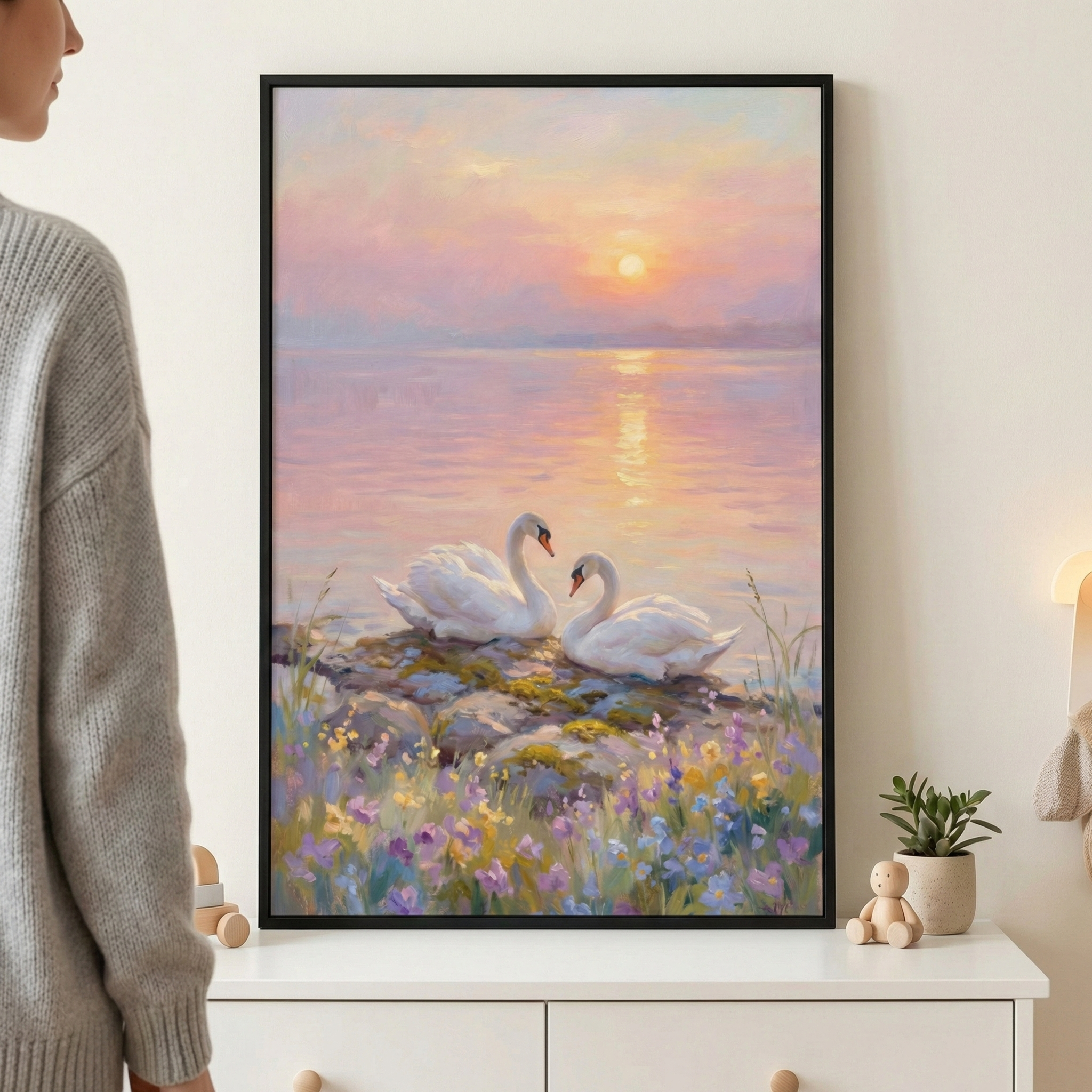 A vertical piece of wall art depicts a tranquil scene of two white swans resting together on a rocky shore at sunset. The sky above the calm water is a soft gradient of pink, orange, and light purple, with a glowing sun reflected on the water's surface. In the foreground, small purple and yellow wildflowers grow among the mossy rocks. The artwork has a soft, impressionistic style with visible textures. It is displayed in a black frame on a white surface, accompanied by a small green plant and a wooden toy.