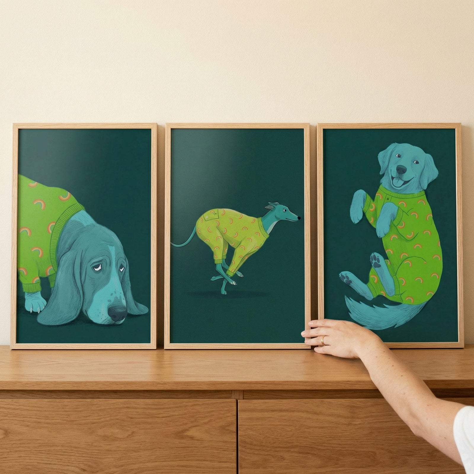 Three vertically oriented, framed illustrations of stylized blue dogs are arranged side-by-side on a wooden dresser. Each illustration features a different breed against a deep teal-green background, and all the dogs are dressed in bright green or yellow-green sweaters with a small, pink, crescent-shaped pattern.
