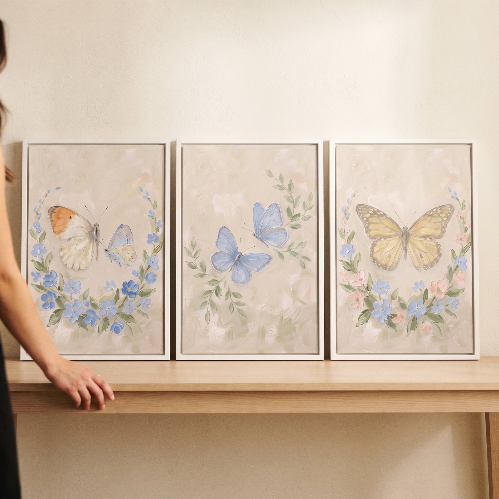 Pale palette garden butterfly wall art set of 3 white framed pieces resting on a light wood table with a hand nearby.