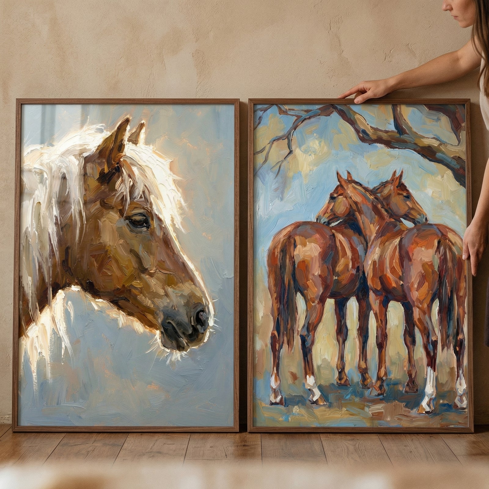 Two framed textured horse paintings displayed side by side, one showing a close up of a brown horse with a light mane against a soft blue background and the other showing two horses standing close together under a tree. A hand adjusts the artwork, suggesting styling in a warm rustic home setting.