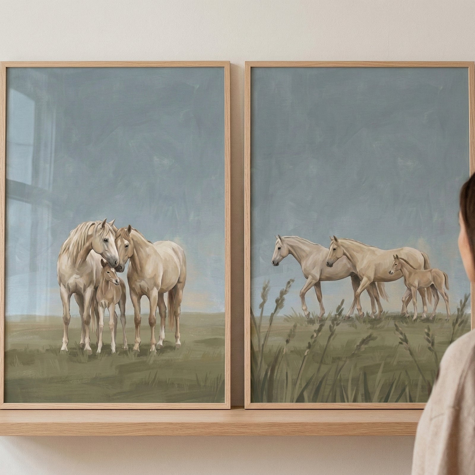 Two wooden frames rest on a shelf against a neutral wall. Each frame showcases an illustration of light-tan horses in a soft, open landscape. The left image features three horses, including a foal, gathered together in a grassy field under a pale blue sky. The right image shows three horses walking in profile through tall blades of grass. The art has a gentle, hand-rendered feel with muted tones. The edge of a person's head and shoulder is visible on the right as they view the artwork.