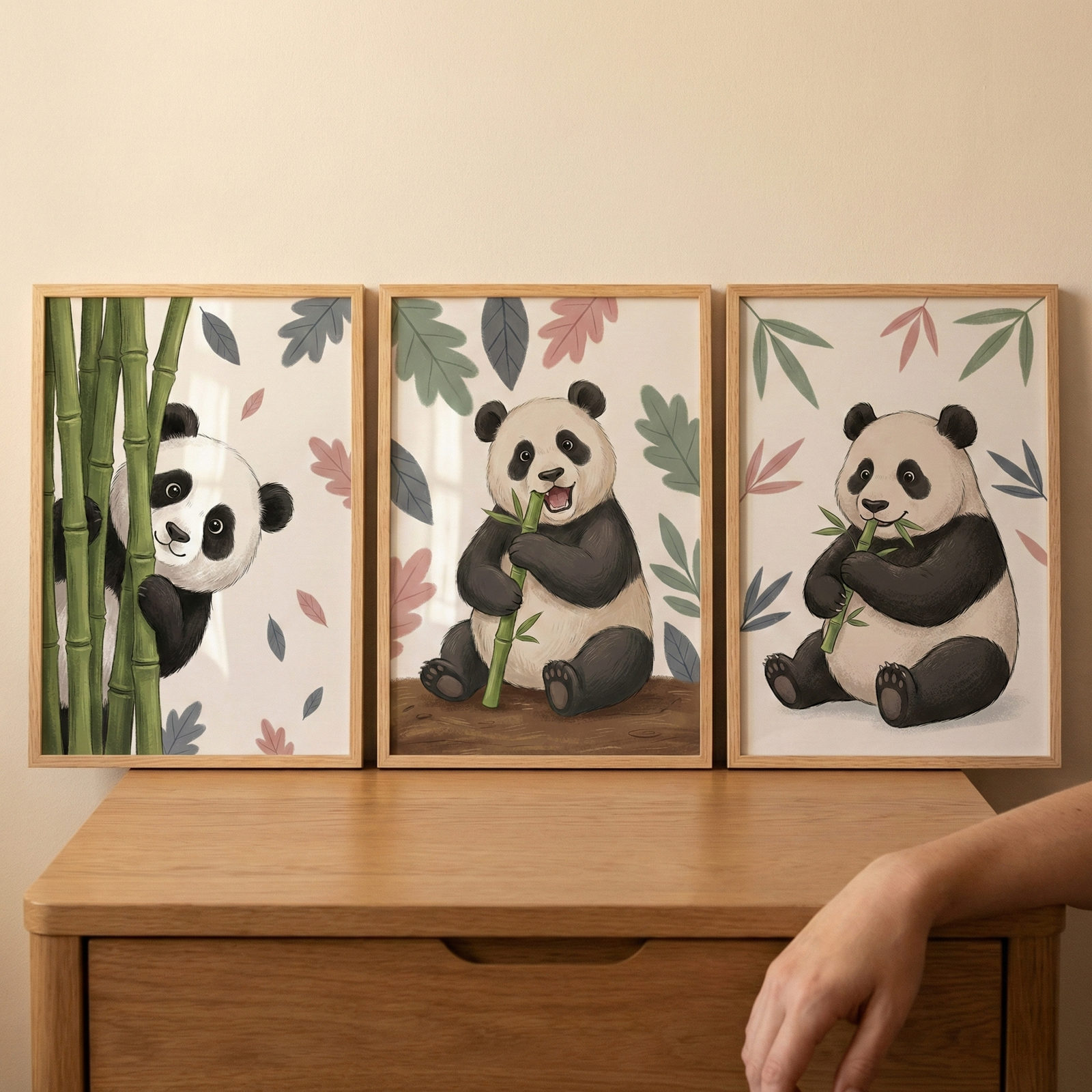 Three framed illustrations of pandas sit side-by-side on a light wood dresser. In the left frame, a panda peeks from behind tall green bamboo. The middle frame features a panda sitting and holding a bamboo stalk. The right frame shows a panda eating bamboo leaves. Each piece has a cream background with soft-colored, scattered leaves. The wooden frames match the surface they rest on. A person's hand is visible on the right side of the wooden furniture.