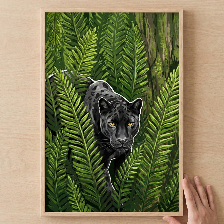A stylized illustration of a black big cat with subtle spots and glowing golden eyes, peeking through a dense thicket of green fern fronds. The animal's form is accentuated with a thin white outline. The artwork is presented in a light wood frame, held by a hand against a wooden surface.