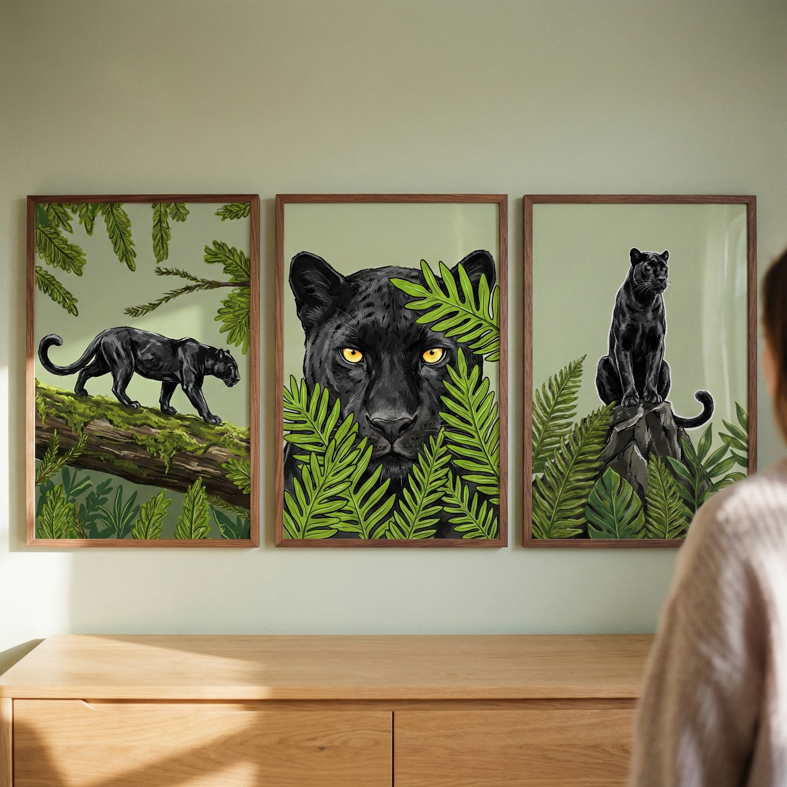 Three framed vertical illustrations hang side-by-side on a light sage green wall above a light-colored wooden dresser. The artwork features a sleek black feline in a tropical jungle setting. On the left, the animal is shown in profile, prowling across a moss-covered tree branch. The center frame is a close-up of the feline's face with bright yellow eyes peering through vibrant green fern leaves. On the right, the creature sits upright on a rock, looking off to the side amidst lush foliage. 
