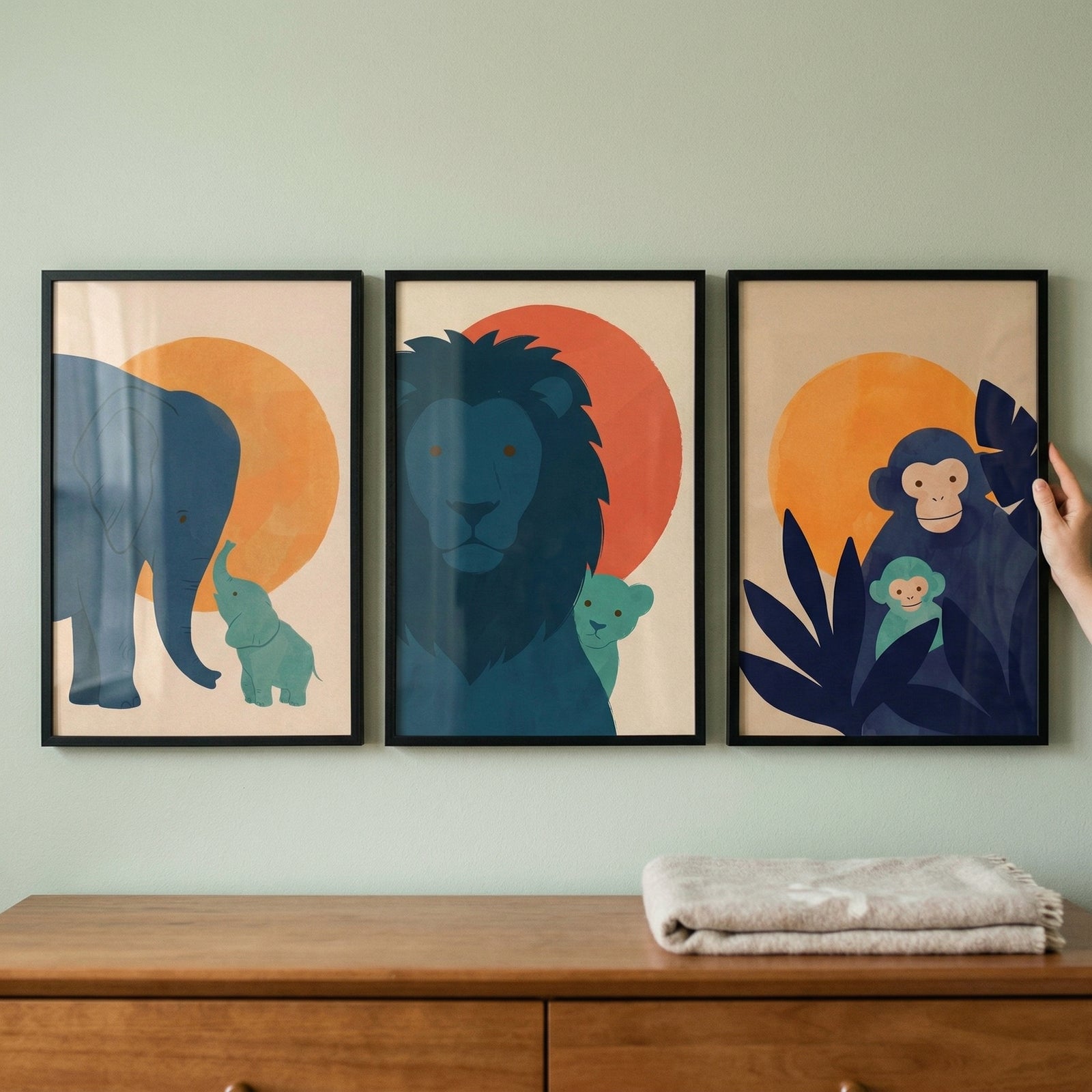 Three framed minimalist art prints of animals and their offspring are displayed on a light green wall. The left print shows a blue elephant and a teal calf. The center print features a blue lion and a small teal cub. The right print depicts two blue primates amidst tropical leaves. 