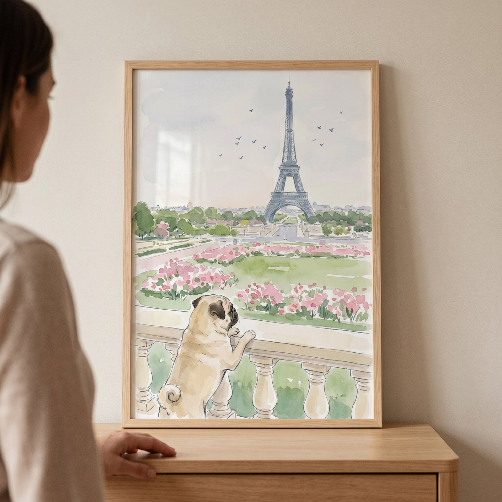 Woman stands by framed paris sightseeing watercolor pug wall art of a pug dog looking at the Eiffel Tower.