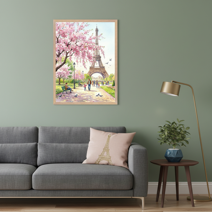 Living room with a gray sectional sofa. A framed watercolor painting of the Eiffel Tower and pink cherry blossom trees in a park hangs above the left side of the sofa. A pink pillow with an Eiffel Tower drawing is on the sofa, with a plant on a side table and a gold floor lamp to the right.