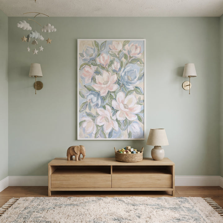 Pastel blue white magnolia floral wall art hangs above a wood console in a sage green room with a lamp and cloud mobile.