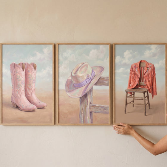 Pastel blush western gear wall art set of 3 with pink boots, a hat on a fence, and a jacket on a wooden chair.