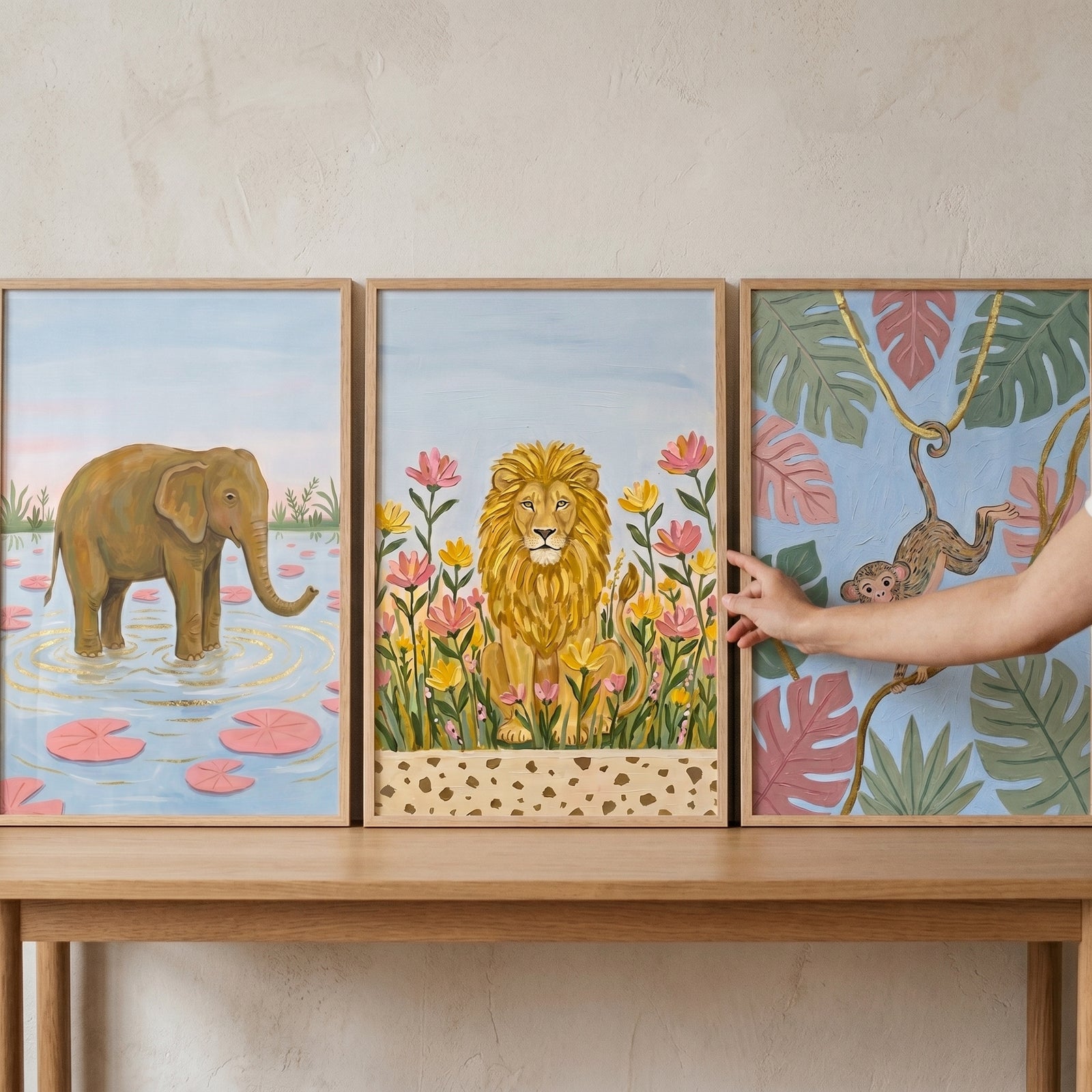 Hand adjusts a pastel botanical safari animals wall art set of 3 featuring an elephant, lion, and monkey on a wooden table.