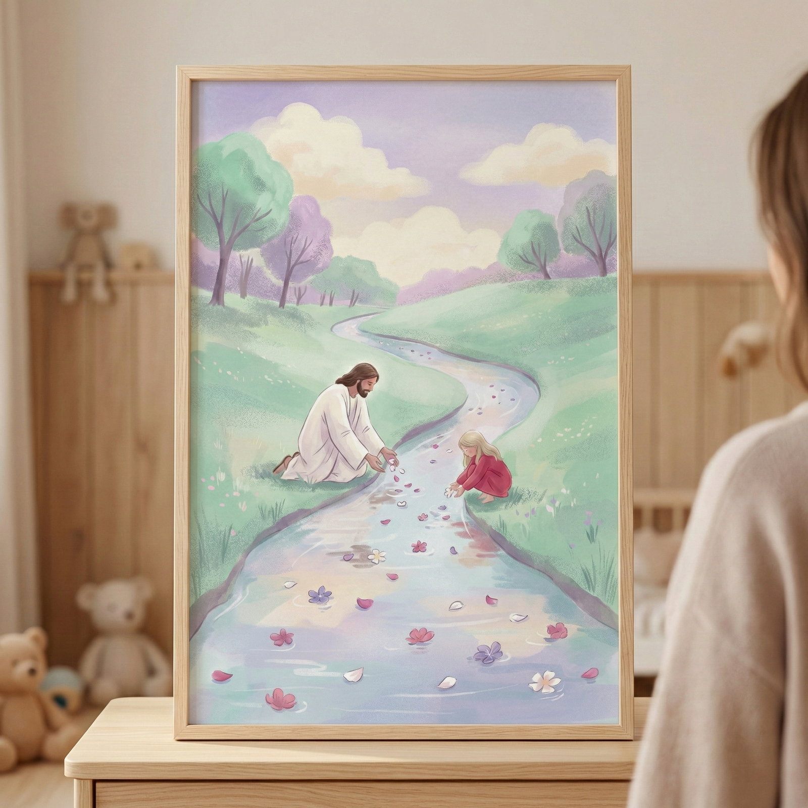 A vertical framed illustration in a light wood frame sits on a wooden surface. The artwork depicts Jesus, dressed in white robes, kneeling by a winding stream alongside a small child in a red dress. Both are gently placing pink and purple flower petals into the water, which float toward the foreground. The serene landscape features rolling green hills and trees in soft pastel shades of lavender and green under a pale, hazy sky. 