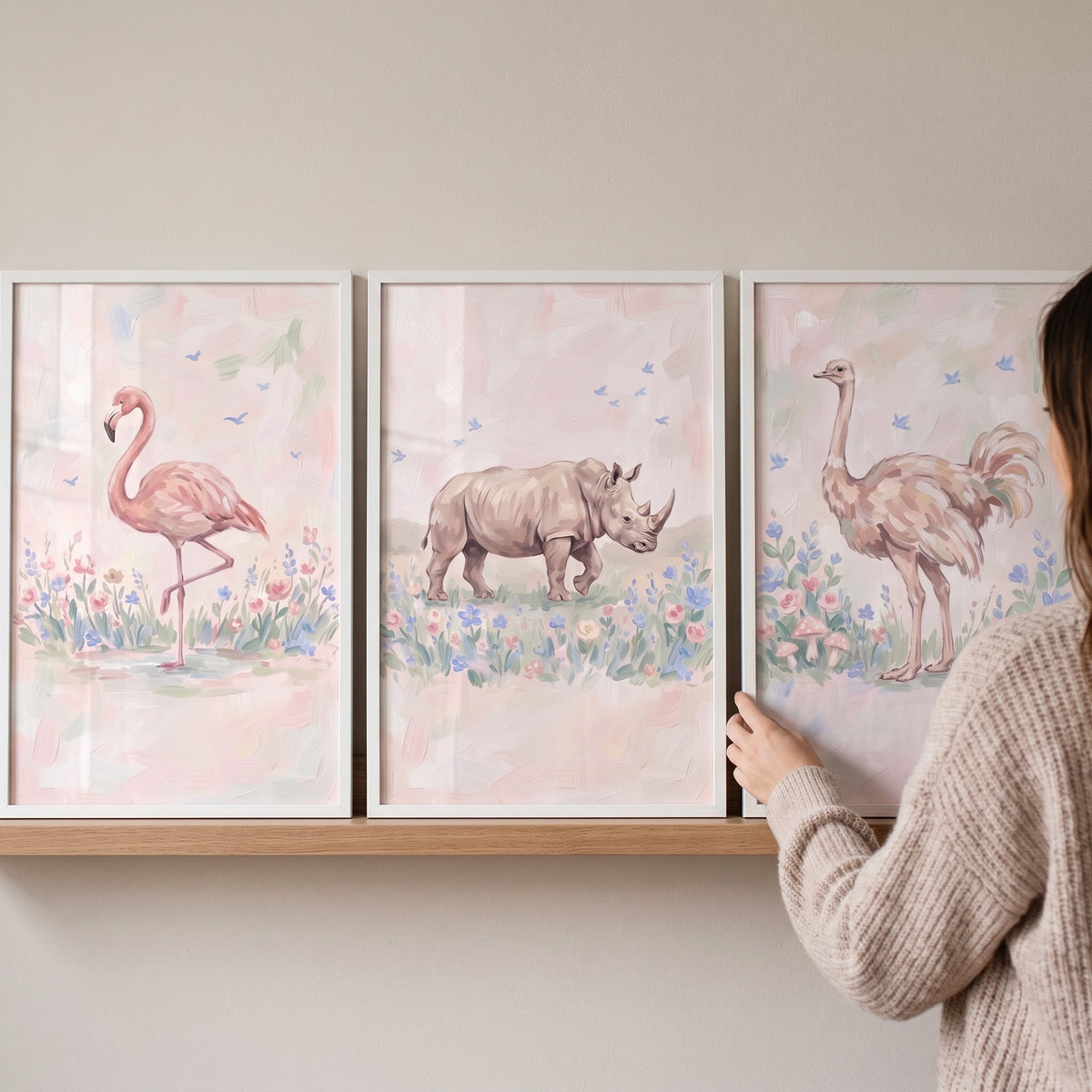 A woman hangs a pastel meadow safari animals wall art set of 3 featuring a flamingo, rhino, and ostrich in white frames.
