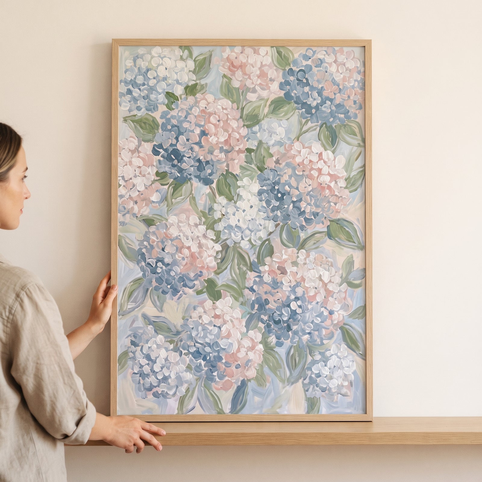 A woman holds a framed pastel pink blue hydrangea floral wall art piece featuring soft flower clusters and green leaves.