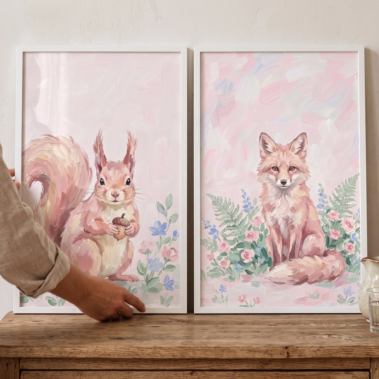 Hand adjusts a pastel pink meadow animals wall art set of 2 with a squirrel and fox in soft floral scenes.