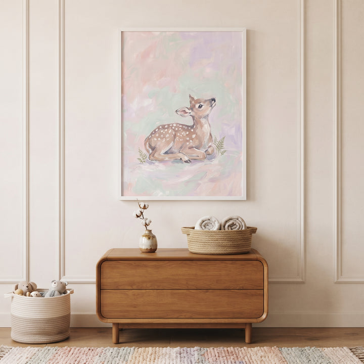 Pastel pink resting fawn wall art hangs above a wooden dresser in a neutral nursery with toys and a striped rug.