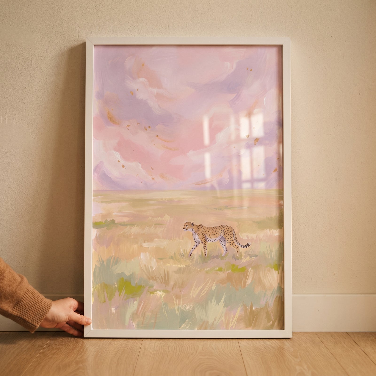 A hand holds a framed pastel pink sky cheetah animal wall art piece of a cheetah walking through a golden field.