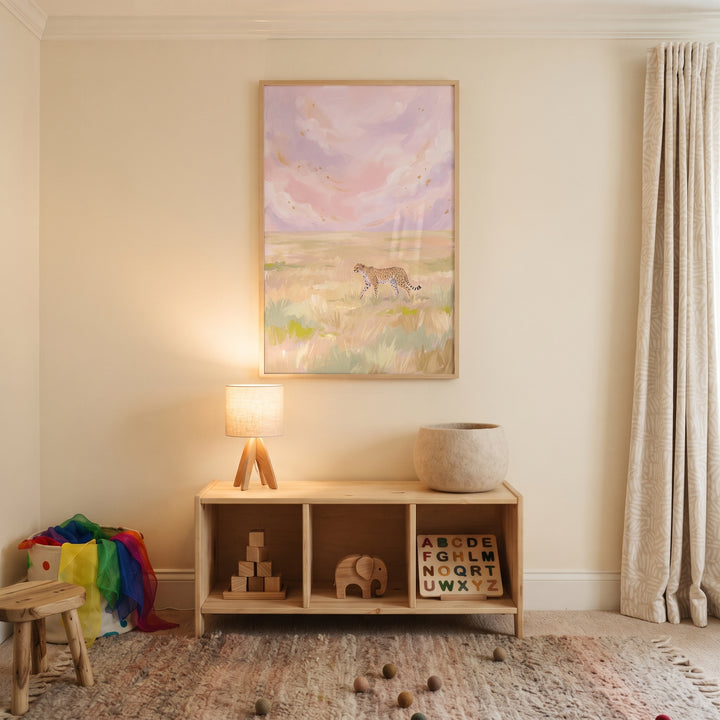 Pastel pink sky cheetah animal wall art hangs above a wooden toy shelf, tripod lamp, and rug in a neutral-toned child's room.
