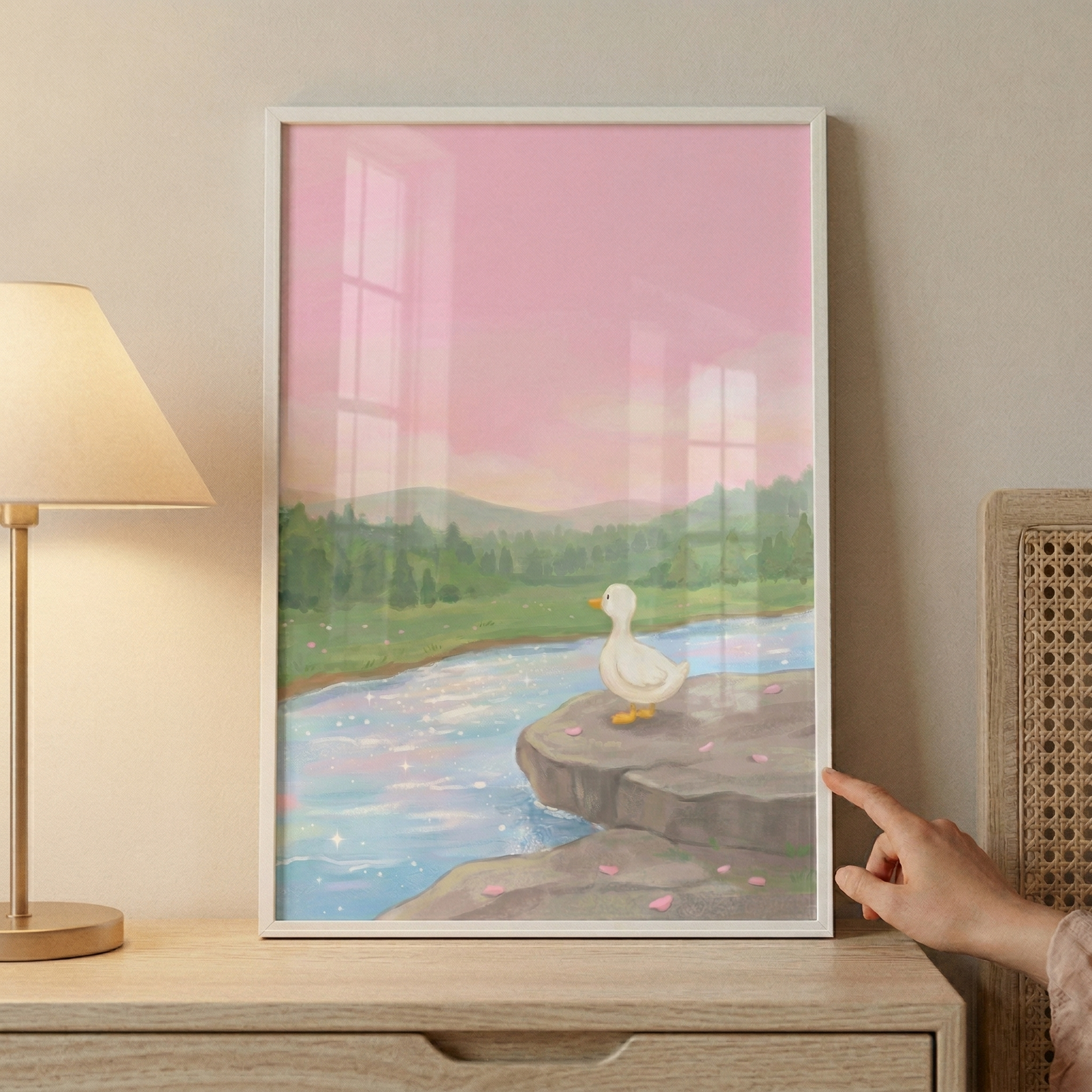 An illustration within a simple white frame depicts a tranquil scene. A white duck with an orange beak is perched on a rocky outcrop overlooking a calm, shimmering river. The water reflects the soft pink and purple tones of a hazy sky, with tiny sparkles of light dancing on its surface. In the background, rolling green hills are densely forested with evergreen trees under a glowing pink sky, evoking a sense of peaceful twilight. Small pink petal-like shapes are scattered on the grey rocks in the foreground.