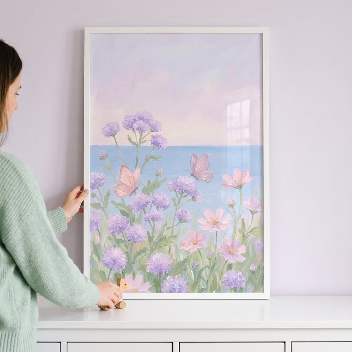 Framed pastel purple floral butterfly wall art being positioned by a person above a white surface.