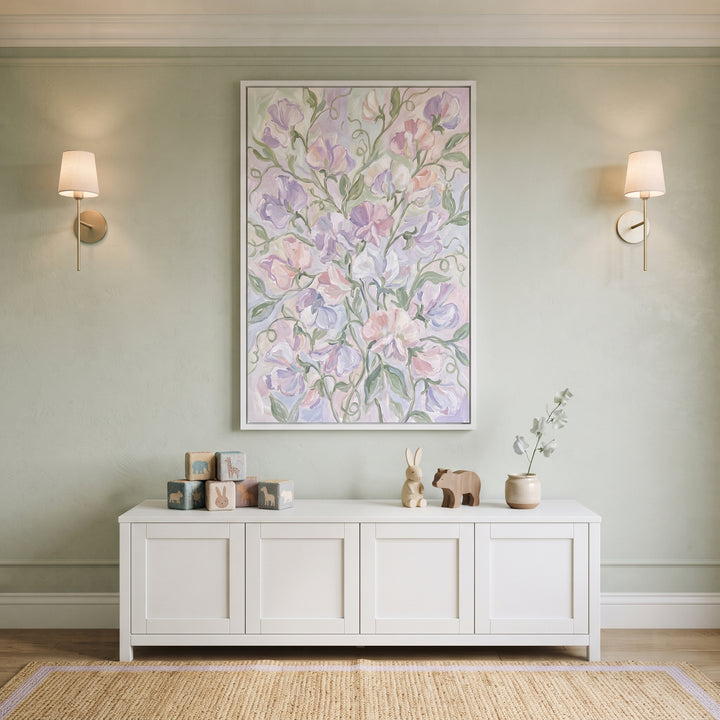 Pastel purple sweet pea floral wall art above a white cabinet with wooden toys in a green room with two sconces.