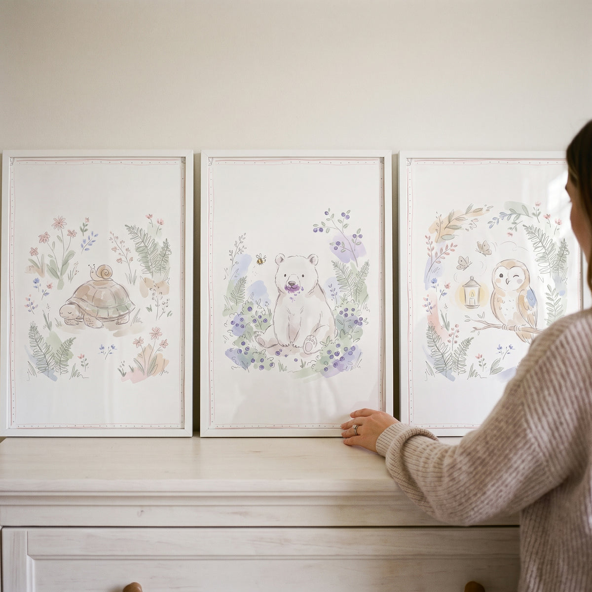 A woman arranges a pastel sketched forest animal wall art set of 3 featuring a turtle, bear, and owl on a white dresser.