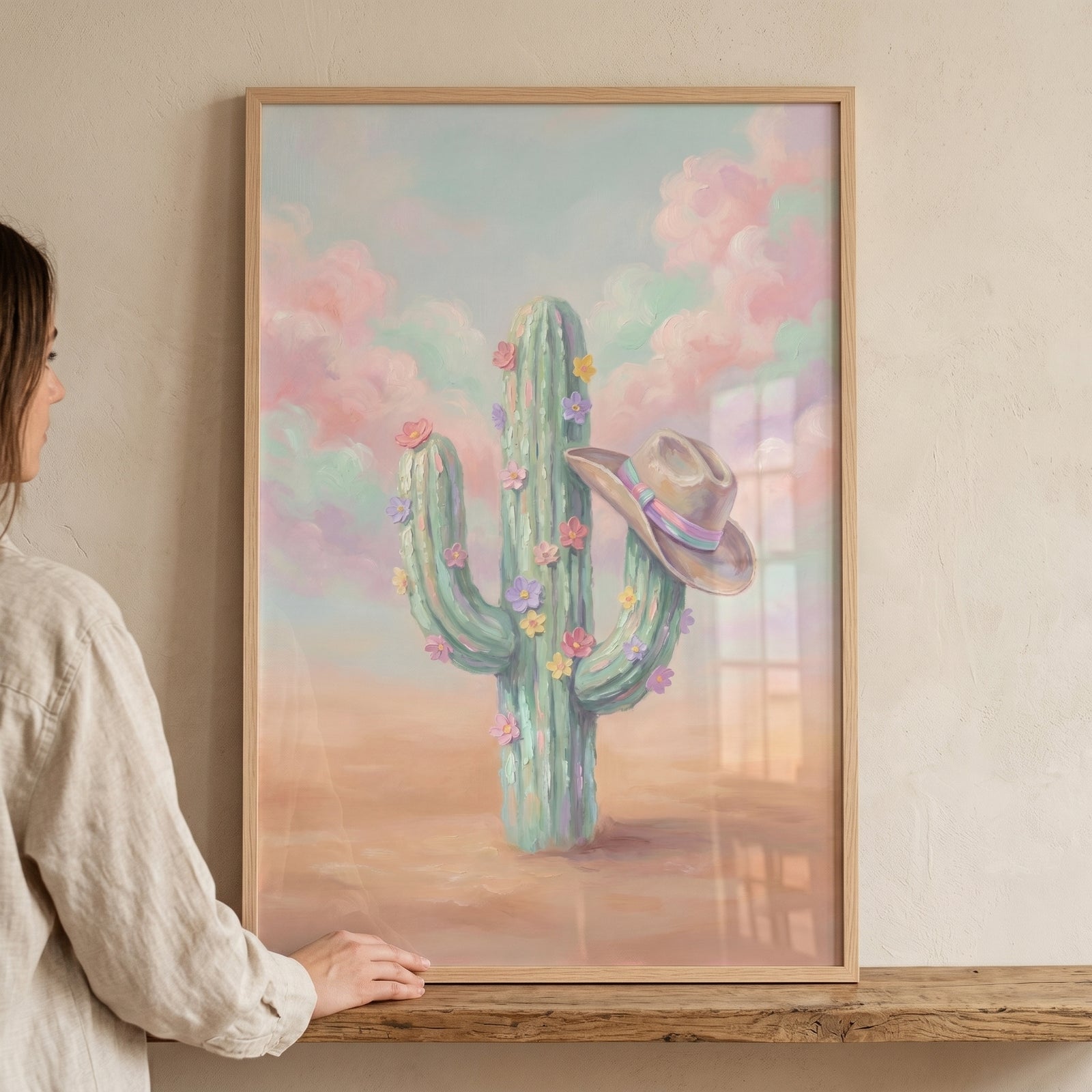 A person holds a framed pastel sky floral cactus wall art featuring a desert cactus with flowers and a cowboy hat.