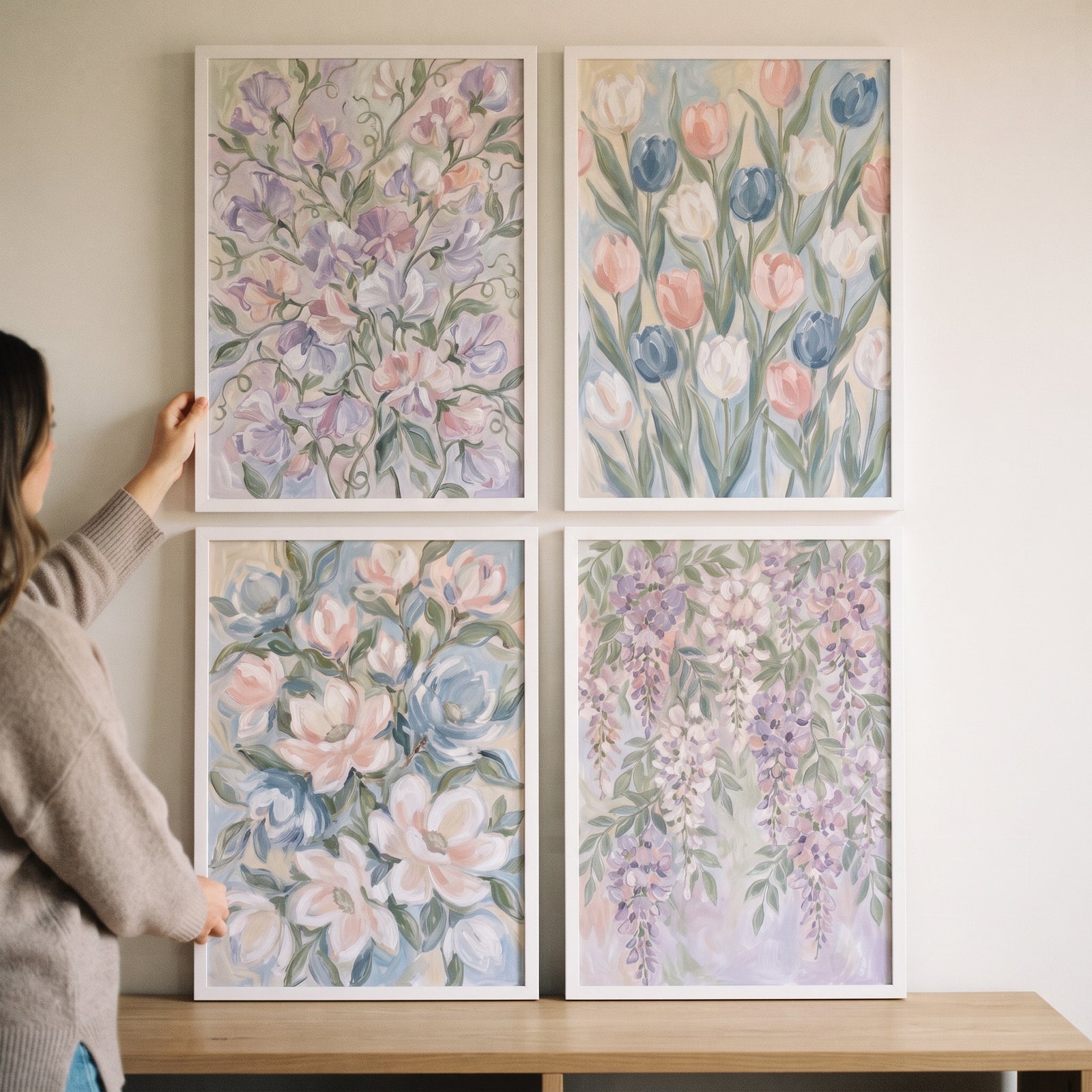 A person hangs a pastel spring garden floral wall art set of 4 in white frames on a wall above a wooden table.