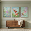 A high-resolution view of a nursery wall featuring three framed vertical artworks arranged in a row. The illustrations are rendered in a soft, whimsical style. The leftmost piece shows a delicate fairy with purple wings and a matching tutu dancing on a lily pad in a pond. The middle artwork depicts two winged figures, one of whom is carrying a large strawberry, flying above a red and white spotted mushroom. The rightmost piece features a fairy in a pink tutu tending to woodland flora. 
