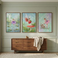 A high-resolution view of a nursery wall featuring three framed vertical artworks arranged in a row. The illustrations are rendered in a soft, whimsical style. The leftmost piece shows a delicate fairy with purple wings and a matching tutu dancing on a lily pad in a pond. The middle artwork depicts two winged figures, one of whom is carrying a large strawberry, flying above a red and white spotted mushroom. The rightmost piece features a fairy in a pink tutu tending to woodland flora. 
